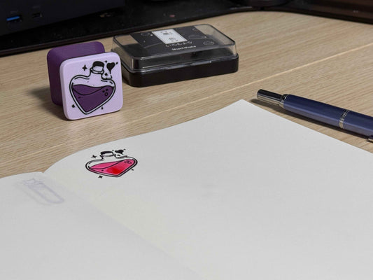 A customized heart-shaped ink stamp stamped on a book with swatched ink, placed on a desk next to a pen and a inkpad.