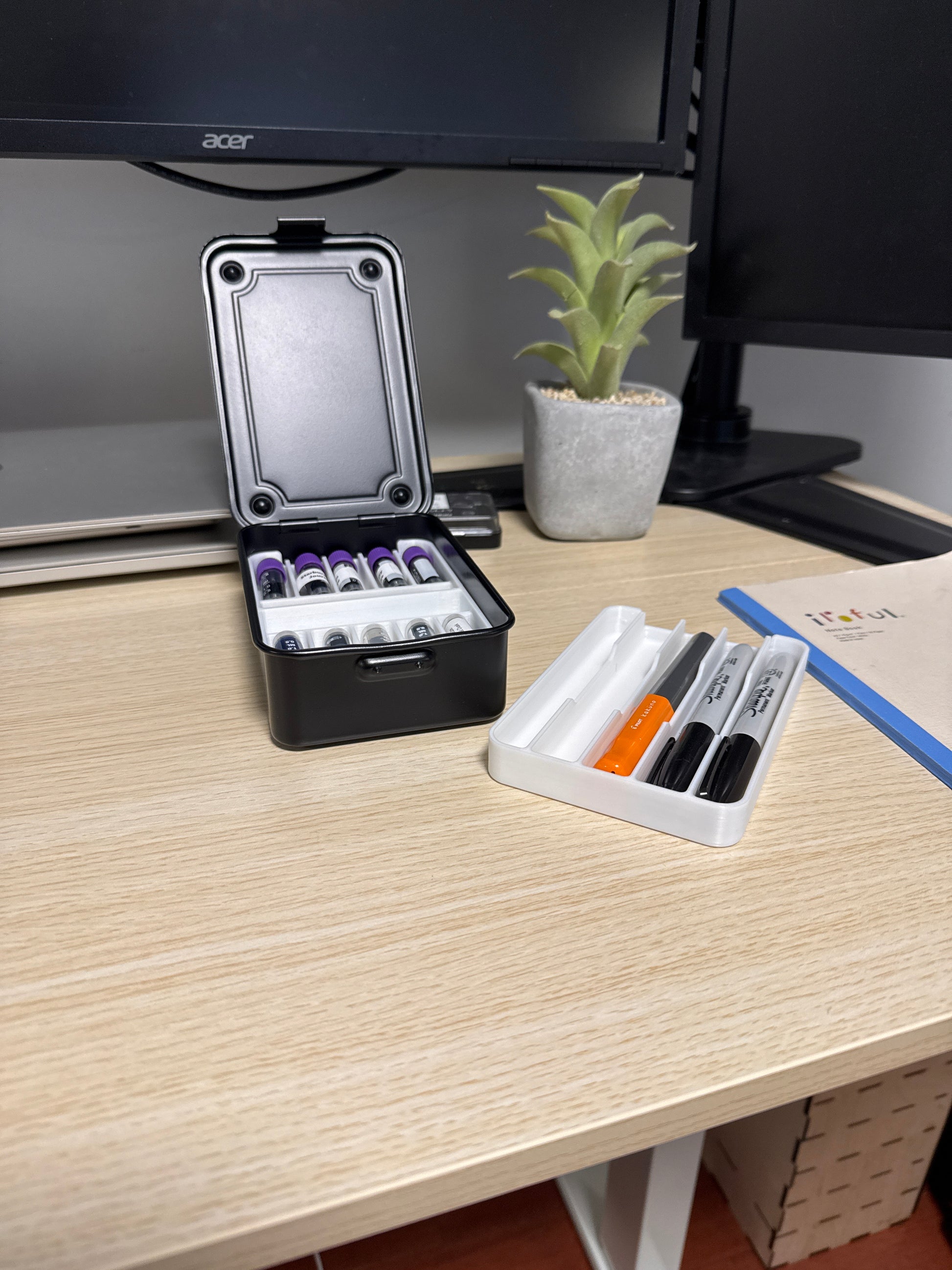 A black Toyo T-150 stackable tool box placed on a wooden surface, with a laptop, plant, and other items in the background. Open with two white inserts.