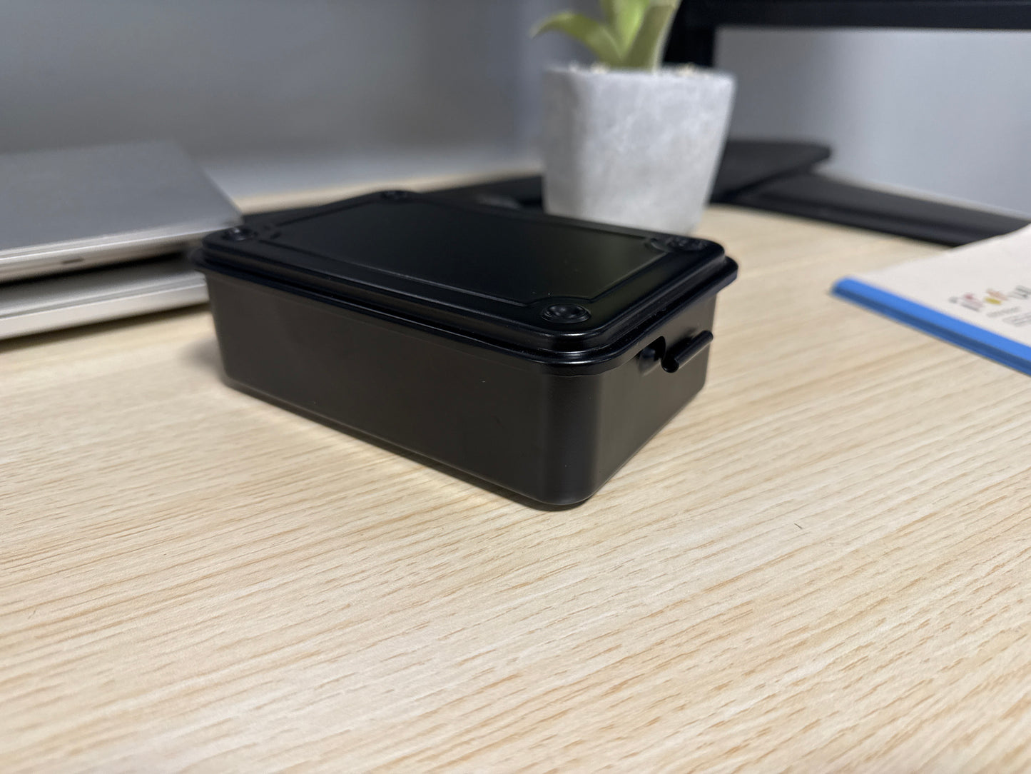A black Toyo T-150 stackable tool box placed on a wooden surface, with a laptop, plant, and other items in the background.